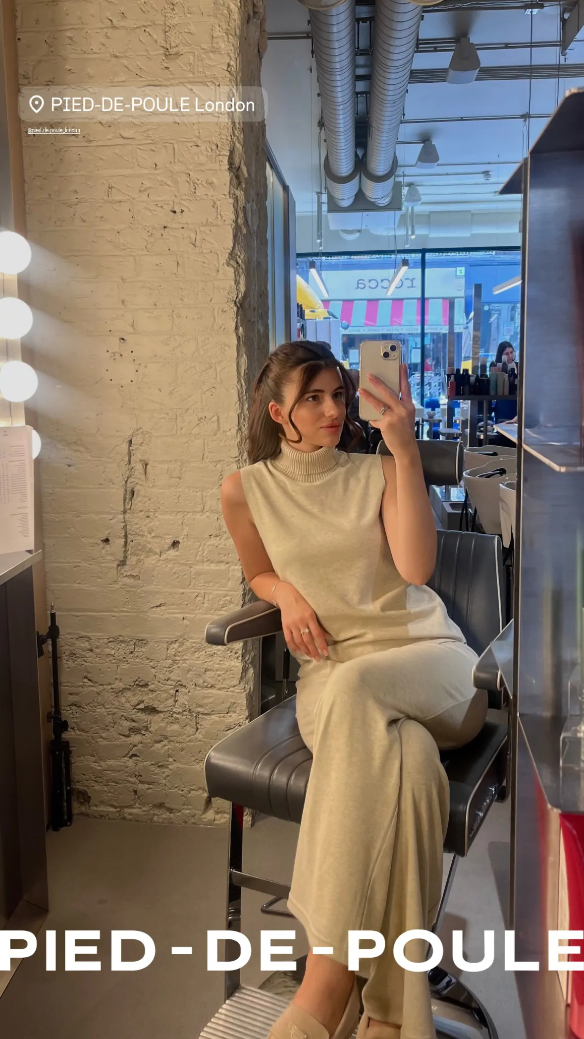 Kobieta robi selfie w odbiciu lustra, prezentując fryzurę i elegancki, beżowy strój w salonie urody „Pied-de-Poule”.